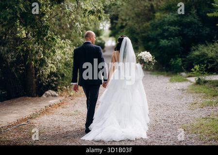 Une mariée aux cheveux noirs dans une robe blanche et un marié dans un costume bleu avec un noeud papillon sur une promenade dans le parc le jour de leur mariage Banque D'Images