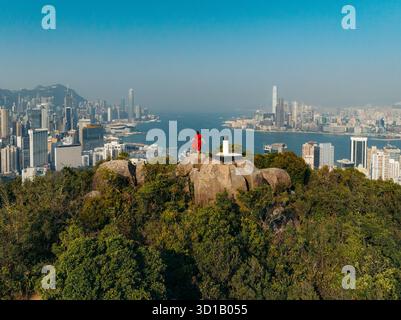 Vue aérienne d'un personnage rouge debout au sommet du Lion Rock, surplombant le paysage urbain tentaculaire et le port Victoria, Hong Kong, Hong Kong. Banque D'Images