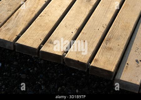Des planches de bois robustes forment un pont de quai, montrant le grain, les nœuds et la texture altérée. Les galets sous ajoutent du contraste, créant une scène extérieure rustique adaptée aux thèmes maritimes ou de construction. Photo de haute qualité Banque D'Images