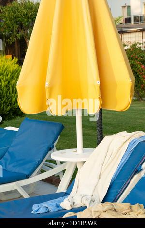 Un centre de villégiature lumineux au bord de la piscine est décoré de grands parasols jaunes, de chaises longues bleues et de palmiers luxuriants. Atmosphère relaxante et ensoleillée parfaite pour les vacances, les loisirs et les expériences de loisirs en plein air. Photo de haute qualité Banque D'Images