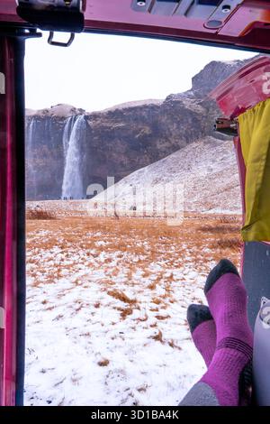 Voyageur se relaxant avec des chaussettes chaudes en regardant la cascade seljalandsfoss et le paysage enneigé de l'intérieur d'un camping-car Banque D'Images