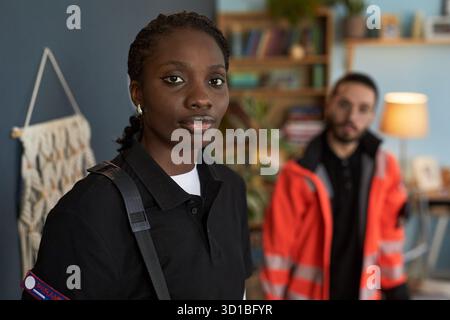 Portrait d'une jeune femme noire adulte debout à l'intérieur portant l'uniforme, regardant dans la caméra avec une expression sérieuse, collègue masculin en arrière-plan portant la veste d'intervention d'urgence, pendant l'appel à domicile Banque D'Images