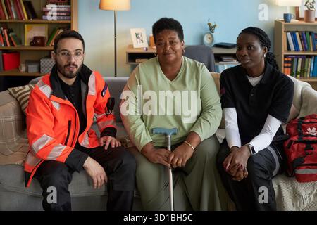 Portrait d'une femme noire d'âge moyen handicapée assise sur un canapé tenant une canne, flanquée d'un jeune homme caucasien adulte et d'une jeune femme noire adulte portant des uniformes paramédicaux Banque D'Images