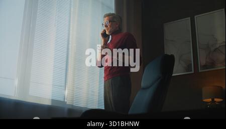 Homme mûr dans le col roulé rouge et les lunettes se tient par une grande fenêtre, boire du café ou du thé tout en étant engagé dans l'appel téléphonique dans la chambre d'hôtel moderne. Concept Business communication, vie professionnelle à distance. Portable. Banque D'Images