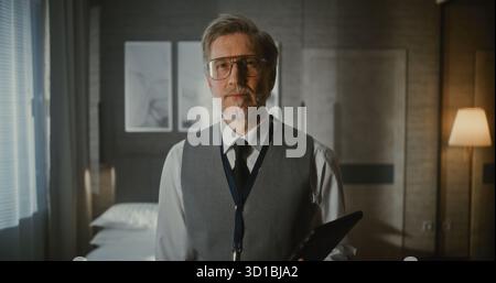 Portrait de Executive dans une chambre d'hôtel moderne. Homme mûr dans Business Vest et cravate se tient avec une expression sérieuse et confiante avec Gadget dans les mains. Concept de exécutif moderne, professionnalisme des affaires. Banque D'Images