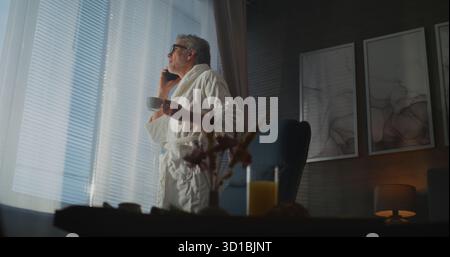 Man in White Hotel peignoir se tient près de la fenêtre, sirotant le thé tout en étant engagé dans l'appel téléphonique, sur le plateau de petit déjeuner de table et verre de jus. Rester professionnellement connecté depuis Comfort of Hotel. Portable. Banque D'Images