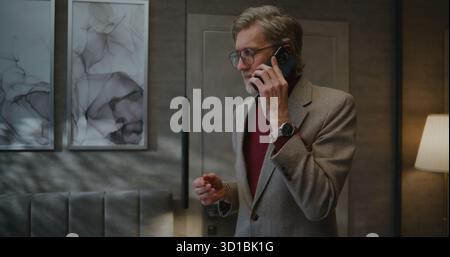 Élégant homme d'affaires européen entre dans la chambre d'hôtel lumineuse avec bagages. Homme menant des négociations commerciales sur smartphone. Concept, voyage d'affaires, vie hôtelière, communication d'affaires, style de vie d'affaires. Banque D'Images