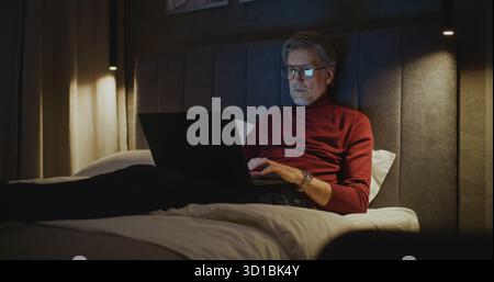 Élégant homme mature portant des lunettes utilise un ordinateur portable tout en étant assis sur le lit. Dans une ambiance chaleureuse de lumière du soir et calme, l'homme travaille sur son ordinateur personnel. Concept Travel, connectivité numérique. Dolly Shot. Banque D'Images