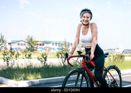 Femme souriante sportive dans le vélo rouge de haut blanc sur la journée ensoleillée près de la zone industrielle du port Banque D'Images