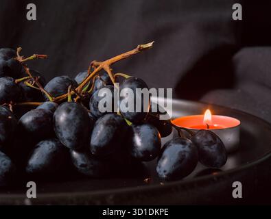 un grand bouquet de raisins mûrs sombres se trouve sur une assiette, illuminé par la lumière chaude de plusieurs bougies allumées Banque D'Images