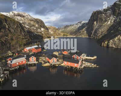 Vue aérienne de charmantes cabanes rouges accrochées aux eaux sombres sous les sommets enneigés, Reine, Nordland, Norvège. Banque D'Images