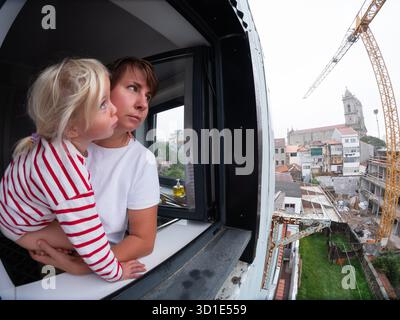 Femme tenant son enfant tout en regardant par la fenêtre, fatiguée du bruit de construction à l'extérieur Banque D'Images