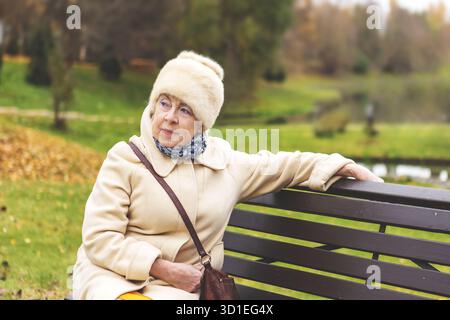 Femme aînée réfléchie assise sur le banc du parc entouré par la nature automnale. Femme âgée appréciant le vieillissement en plein air, paisible et la beauté naturelle à l'automne Banque D'Images