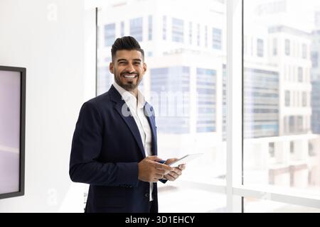 Joyeux jeune homme d'affaires indien dans élégant costume formel tenant la tablette Banque D'Images