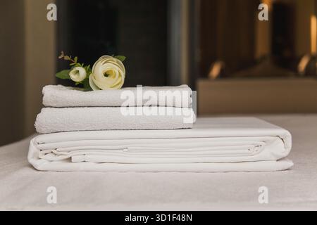 Des serviettes blanches et un drap plié sont placés soigneusement sur un lit. Une petite fleur décore la configuration, lui donnant un aspect frais et invitant. Le paramètre Banque D'Images