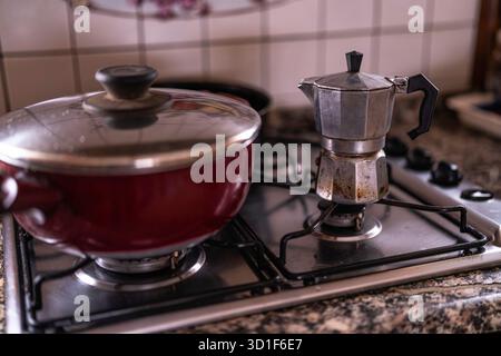 Pot de moka traditionnel à côté d'un pot de cuisson rouge sur une cuisinière à gaz au-dessus du comptoir de granit, la vapeur montant comme le café de petit déjeuner et le repas du matin sont préparés i. Banque D'Images