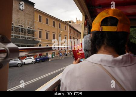 Vue sur la rue depuis une fenêtre de bus touristique de Rome, avec des bâtiments ocres historiques, des voitures garées et un touriste tenant un guide « Big bus Tours ». Banque D'Images