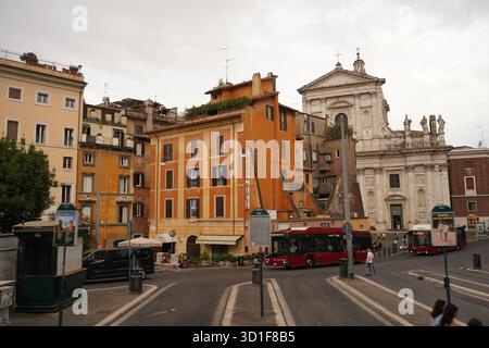 Une intersection romaine animée avec des bus rouges de transport en commun qui passent, une façade d'église baroque blanche et des immeubles d'appartements historiques animés. Banque D'Images
