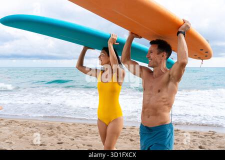 Couple marchant sur une plage de sable, portant des planches de surf sur les épaules, prêt pour un style de vie actif et relever le défi d'apprendre une nouvelle eau Banque D'Images
