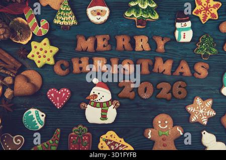 Des biscuits de Noël colorés en forme de bonhommes de neige, d'arbres, d'étoiles et de pain d'épice entourent le texte Joyeux Noël 2026 sur un fond en bois. Banque D'Images