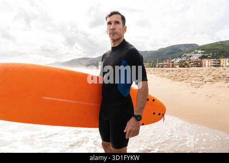 Homme portant une combinaison debout sur la plage de sable, tenant une planche de surf, anticipant une session de surf avec des vagues se brisant sur le rivage et les montagnes dans le Banque D'Images