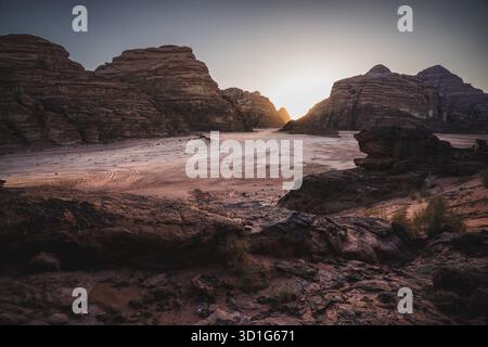 La lumière du coucher du soleil tombe sur les falaises de grès et le sol rouge du désert de Wadi Rum dans le sud de la Jordanie. Un petit camp bédouin et un véhicule sont assis à la base. Banque D'Images