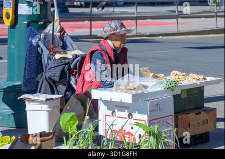 Un marchand de rue âgé portant un chapeau vend du gingembre et des légumes dans des boîtes sur un trottoir Chinatown. Banque D'Images