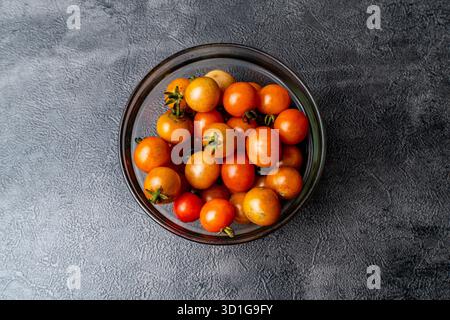 Vue de dessus plat de tomates cerises fraîches dans un bol en verre. Le bol repose sur un fond d'ardoise sombre et texturé. Banque D'Images