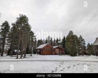 Une belle scène hivernale avec une cabane en bois rustique entourée d'un sol enneigé et de grands pins sous un ciel nuageux. Banque D'Images