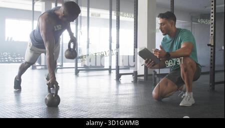 Homme de rangée renégat exécutant tenant deux kettlebells au gymnase avec tablette d'entraîneur, espace de copie Banque D'Images