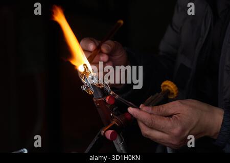 Artiste souffleur de verre démontrant la technique de la flamme de la torche, cadre d'atelier nocturne, personne dans la veste sombre actionnant la torche de verre, pratique artistique de Banque D'Images