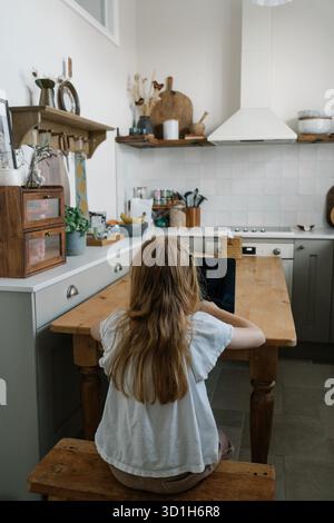 Un enfant assis à une table de cuisine en bois, regardant une tablette dans un intérieur confortable et chaleureux. Banque D'Images