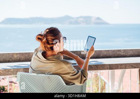 Belle femme d'âge moyen se relaxant avec e-book sur la terrasse ensoleillée de la maison éclairée par la lumière du soleil du matin, surplombant le port de la mer Méditerranée dans la vieille ville de Kas, Banque D'Images