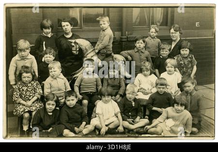 Carte postale originale et charmante du début des années 1900 d'enfants d'écoles maternelles posant dans la rue à l'extérieur d'une école primaire urbaine - peut-être un quartier pauvre, avec des enseignants. Un garçon est assis sur un cheval à bascule. Circa 1925, Royaume-Uni Banque D'Images