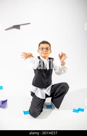 Un garçon joyeux d'origine moyen-orientale jette ludique un avion en papier dans un studio, vêtu d'un costume rayé et de lunettes. Banque D'Images