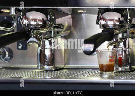 Cremona, Italie - 21 juillet 2025 machine espresso professionnelle en acier inoxydable préparant deux doses de café foncé riche dans de petites tasses en verre sur un occupé Banque D'Images