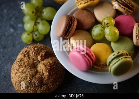 Une nature morte propre montre une petite assiette blanche de macarons colorés avec des raisins et un biscuit sur une table sombre. Le cadrage serré et la lumière douce créent Banque D'Images