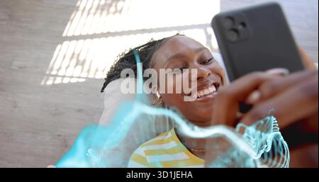 Femme souriante portant une chemise rayée écoutant le téléphone à la maison, avec des écouteurs sans fil et forme d'onde Banque D'Images