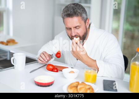 Homme mange le petit déjeuner dans la cuisine le matin. Man 40s aime un repas sain à la table de la cuisine. Un homme mange des céréales et des fruits dans la cuisine pendant Banque D'Images