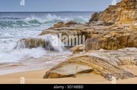 Vague d'eau floue se brisant sur des roches de grès orange et jaune par jour couvert à Maitland Bay, parc national de Bouddi, Nouvelle-Galles du Sud, Australie Banque D'Images
