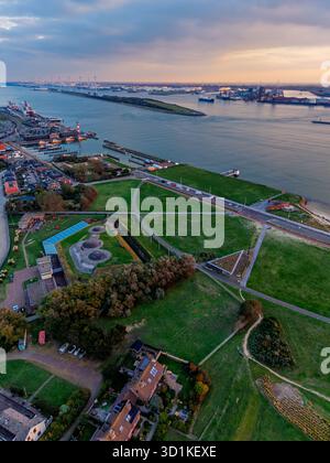 Vue aérienne du port fluvial au coucher du soleil des navires de fret, des grues et des quais industriels bordent les deux rives, avec une grille urbaine dense et des champs verts encadrant l'ouate Banque D'Images