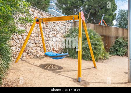 Une seule balançoire jaune et bleue, probablement un siège adapté, se trouve dans une aire de jeux de sable pour enfants contre un mur décoratif en pierre Banque D'Images