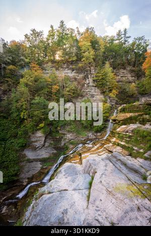 Une longue gorge en gradins montre une fine cascade sur une dalle de roche pâle, des pruches illuminées par le soleil et des arbres teintés par l'automne au parc d'État Robert H. Treman, New York. Banque D'Images