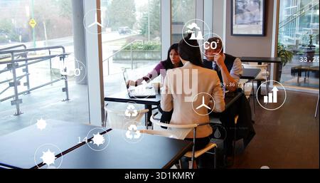 Discussion de trois professionnels analysant les superpositions de données dans le salon de café, avec ordinateur portable et icônes écologiques Banque D'Images
