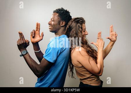 Un jeune homme et une jeune femme se tiennent dos à dos, fabriquant de manière ludique des armes à doigt avec leurs mains. Leurs expressions amicales reflètent un sentiment de plaisir et de camarade Banque D'Images