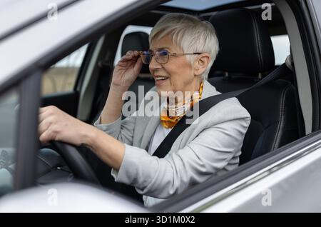 Femme senior en tenue d'affaires conduisant une voiture moderne et ajustant son aveuglement Banque D'Images