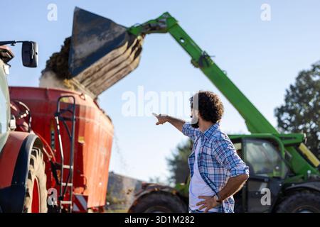 Agriculteur debout devant le tracteur, le mélangeur et le chargeur tout en mélangeant les aliments pour le bétail à la ferme Banque D'Images
