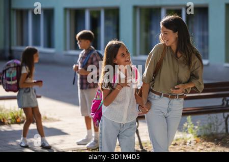 Une mère marche main dans la main avec sa fille heureuse, profitant de leur temps ensemble à l'extérieur de l'école tandis que le garçon marche en arrière-plan Banque D'Images
