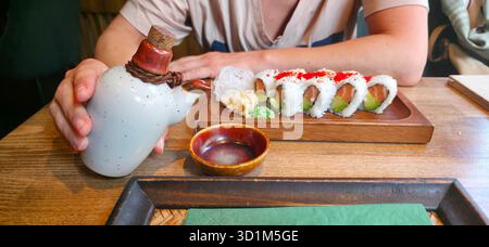 Main tenant une bouteille de saké traditionnelle à côté d'un plateau de sushis magnifiquement arrangé avec wasabi et gingembre mariné, mettant en vedette culinaire Banque D'Images