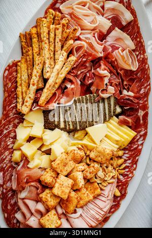 Vue de dessus d'une grande planche de charcuterie avec des tranches de charcuterie salées, des fromages assortis, des arachides et des gressins croustillants. Banque D'Images
